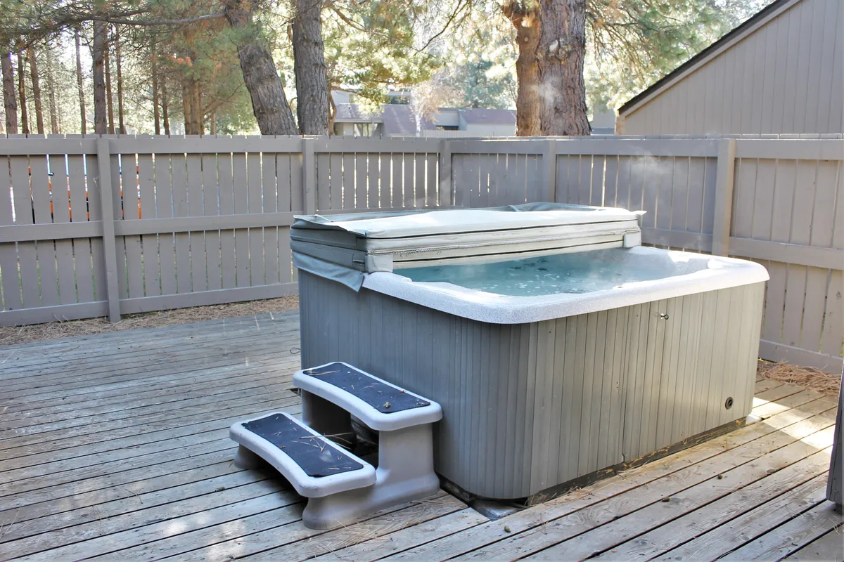 A serene exterior view featuring a private hot tub nestled on a wooden deck, surrounded by mature trees. The tranquil setting highlights both the inviting hot tub and the spaciousness of the deck, with glimpses of nature all around, suggesting privacy and relaxation.