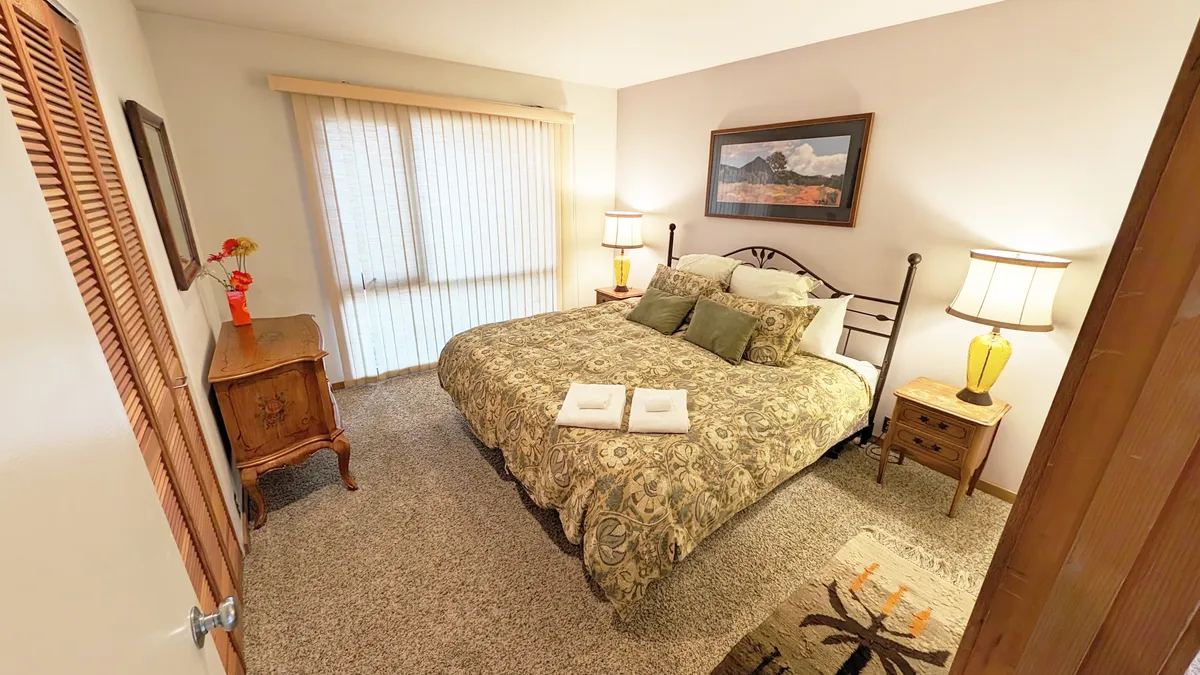 A serene bedroom featuring a large bed adorned with decorative bedding, flanked by nightstands with lamps. Artwork hangs above the bed, and a dresser with decorative flowers sits nearby. The atmosphere is tranquil and filled with soft, natural light, ideal for restful mornings or cozy evenings.