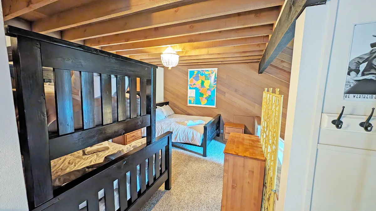 This image displays a loft-style bedroom featuring a queen bed, a pyramid bunk (single over double), a world map, a wooden chest, and a chandelier overhead. The space is bright and partly open, inviting family gatherings and playful activities.