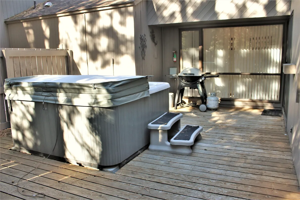 An inviting deck scene with a prominent hot tub, grill, and wooden deck area. The image captures a space ideal for both relaxation and social gatherings, with open views and comfortable outdoor amenities, suggesting a connection to the natural surroundings.