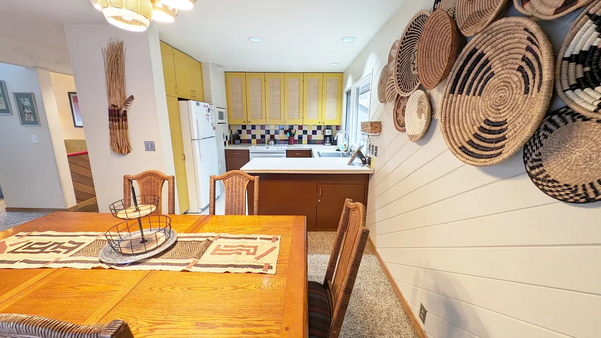 A warm and inviting view of a dining area, with a central dining table, woven wall art, and a visible transition into the kitchen. The setup encourages togetherness for meals and celebrations.