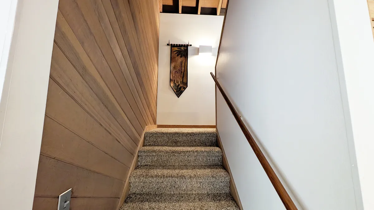 The focal point is a staircase with a decorative wall hanging and wooden railing, symbolizing access to the upstairs loft. The warm, welcoming mood and suggestion of upward movement reinforce the theme of returning to a flexible, relaxing space after outdoor adventures.
