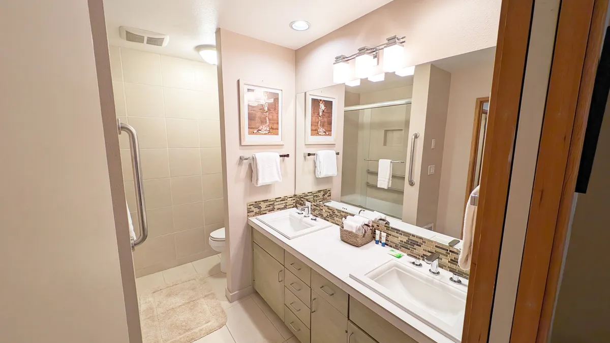 A refreshing, spa-like bathroom featuring a double sink vanity, elegant framed artwork, and a spacious shower area. The atmosphere is bright and serene, perfect for morning routines or relaxing self-care after a day of adventure.