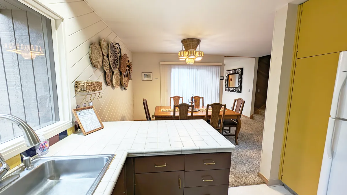 An open kitchen view with a kitchen sink, dining table, decorative wall baskets, and a chandelier. The setting is bright and welcoming, suggesting space for cooking together, dining, and socializing.