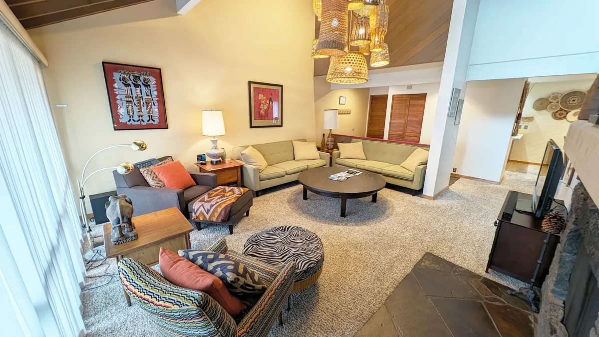 The image showcases a bright, stylish living area with a striking pendant light, colorful accent chairs arranged around a modern coffee table with a decorative tray, and hints of a cozy fireplace. The space exudes warmth and comfort, inviting guests to relax, socialize, and enjoy artistic touches throughout the room.