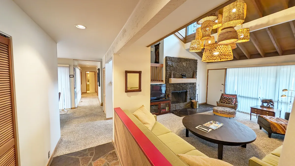 A spacious living room with a large circular coffee table, exposed beam ceiling, stone fireplace, unique pendant lighting, and a comfortable seating area. Sunlight streams in, making the room feel both cozy and bright, perfect for gathering or relaxing fireside.
