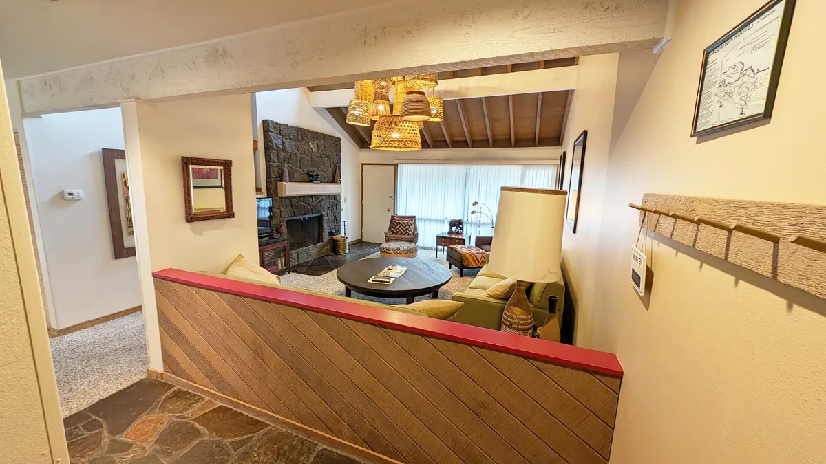 A cozy living area with a stone fireplace, a round coffee table surrounded by comfortable sofa seating, and a large chandelier overhead. Natural light fills the room, and the layout encourages group relaxation and socializing.