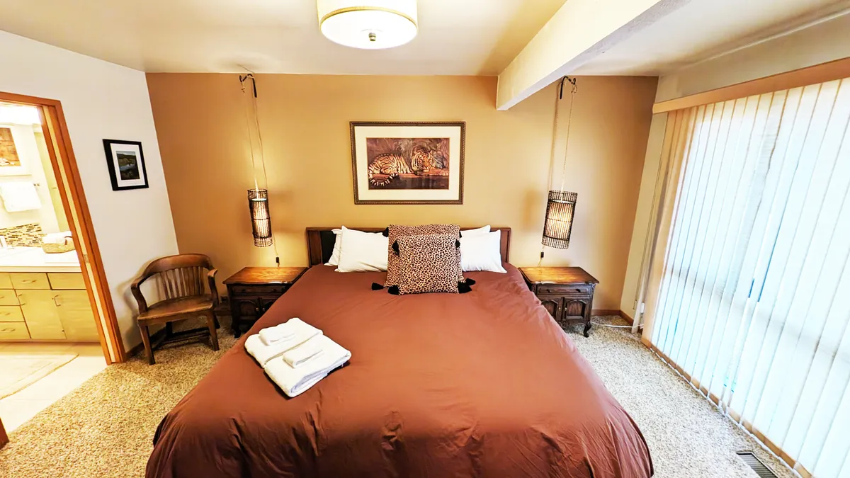An inviting bedroom with a spacious bed, framed artwork, wooden nightstands, hanging light fixtures, and a window with blinds allowing sunlight to fill the room. The décor is warm and modern, creating a peaceful retreat for guests.