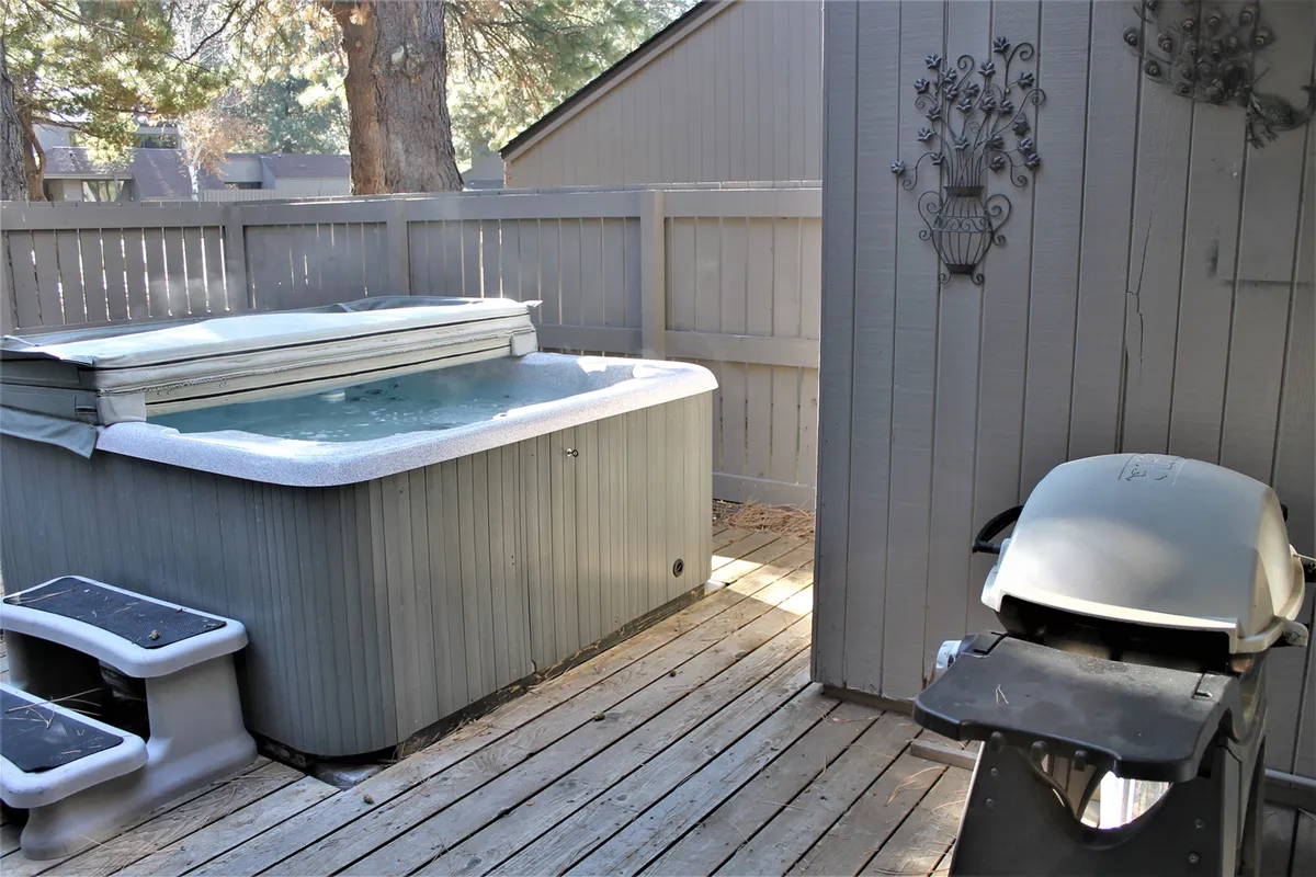 An inviting wooden deck featuring a spacious hot tub and a grill, set against the backdrop of mature trees and tranquil greenery. The area suggests a comfortable space for relaxation and shared meals outdoors.